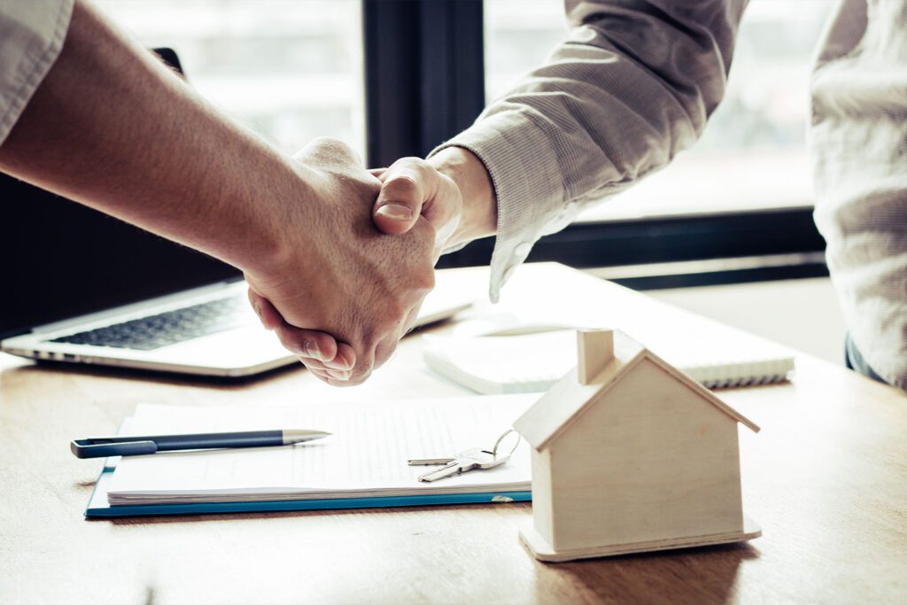 Two people shaking hands over a table with a house on it.